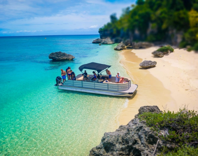 A boat with people is anchored near a sandy beach with turquoise water. Nearby, rocks and lush greenery border the shore, conveying a serene tropical vibe.