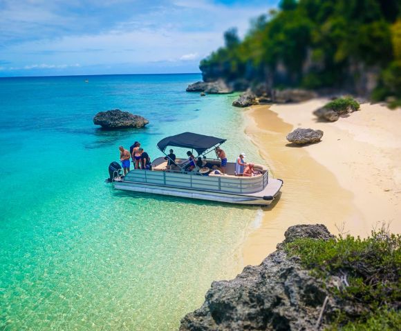 A boat with people is anchored near a sandy beach with turquoise water. Nearby, rocks and lush greenery border the shore, conveying a serene tropical vibe.