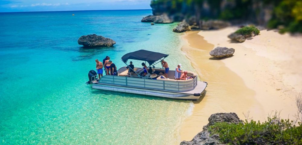 A boat with people is anchored near a sandy beach with turquoise water. Nearby, rocks and lush greenery border the shore, conveying a serene tropical vibe.