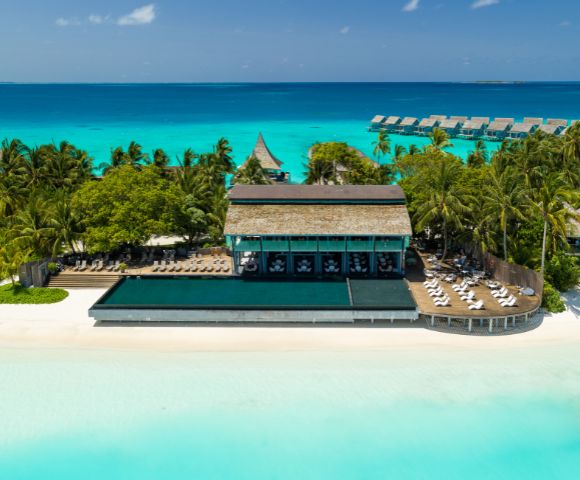 Aerial view of a tropical beach resort with palm trees, a large infinity pool, and sun loungers. Turquoise ocean and overwater bungalows in the background.
