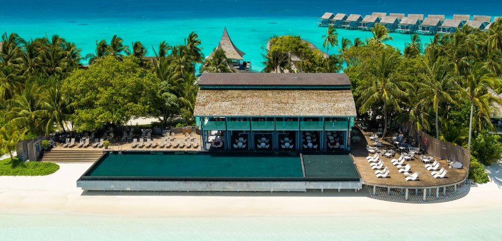 Aerial view of a tropical beach resort with palm trees, a large infinity pool, and sun loungers. Turquoise ocean and overwater bungalows in the background.
