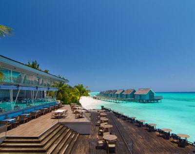 Luxurious beachside resort with modern wooden deck, ocean-view dining area, palm trees, and overwater bungalows. Vibrant turquoise sea under a clear blue sky.