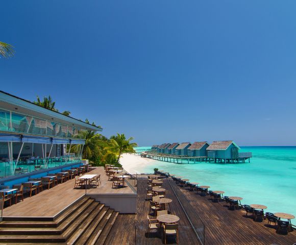 Luxurious beachside resort with modern wooden deck, ocean-view dining area, palm trees, and overwater bungalows. Vibrant turquoise sea under a clear blue sky.