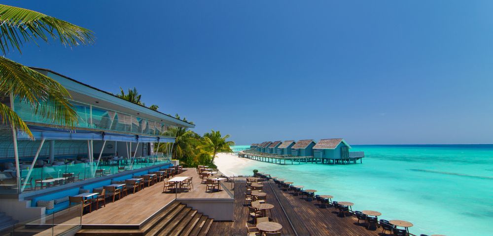 Luxurious beachside resort with modern wooden deck, ocean-view dining area, palm trees, and overwater bungalows. Vibrant turquoise sea under a clear blue sky.