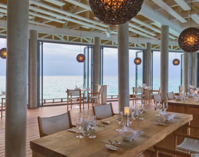 Elegant beachfront restaurant with wooden tables, set with glassware, overlooking the ocean. Warm, spherical pendant lights enhance a serene ambiance.