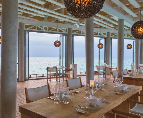Elegant beachfront restaurant with wooden tables, set with glassware, overlooking the ocean. Warm, spherical pendant lights enhance a serene ambiance.