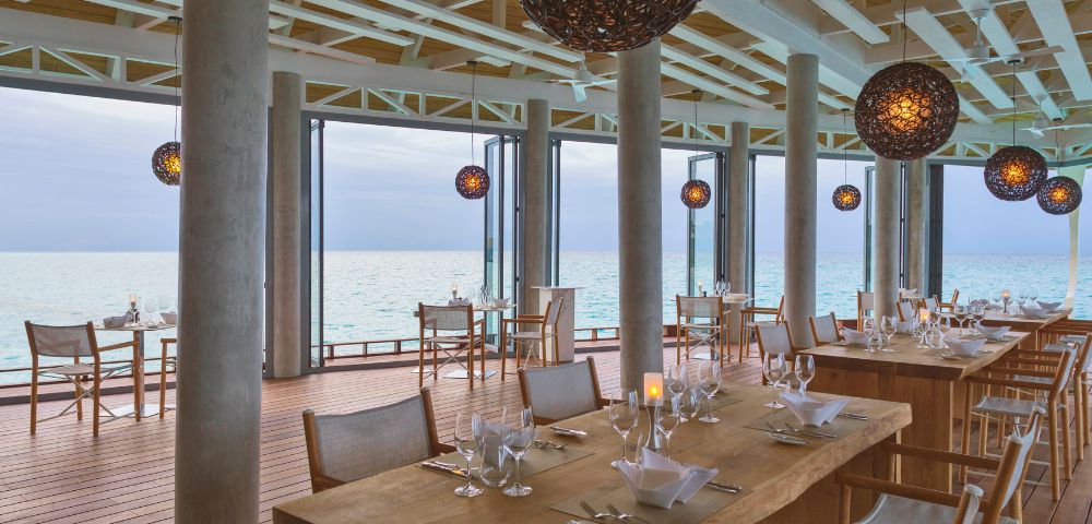 Elegant beachfront restaurant with wooden tables, set with glassware, overlooking the ocean. Warm, spherical pendant lights enhance a serene ambiance.