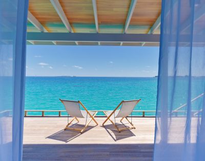 Two deck chairs on a wooden terrace overlook a tranquil turquoise sea under a clear blue sky, framed by sheer curtains, conveying relaxation.