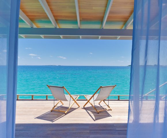 Two deck chairs on a wooden terrace overlook a tranquil turquoise sea under a clear blue sky, framed by sheer curtains, conveying relaxation.