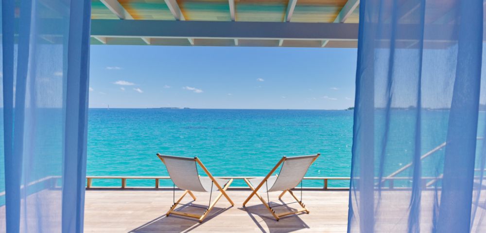 Two deck chairs on a wooden terrace overlook a tranquil turquoise sea under a clear blue sky, framed by sheer curtains, conveying relaxation.