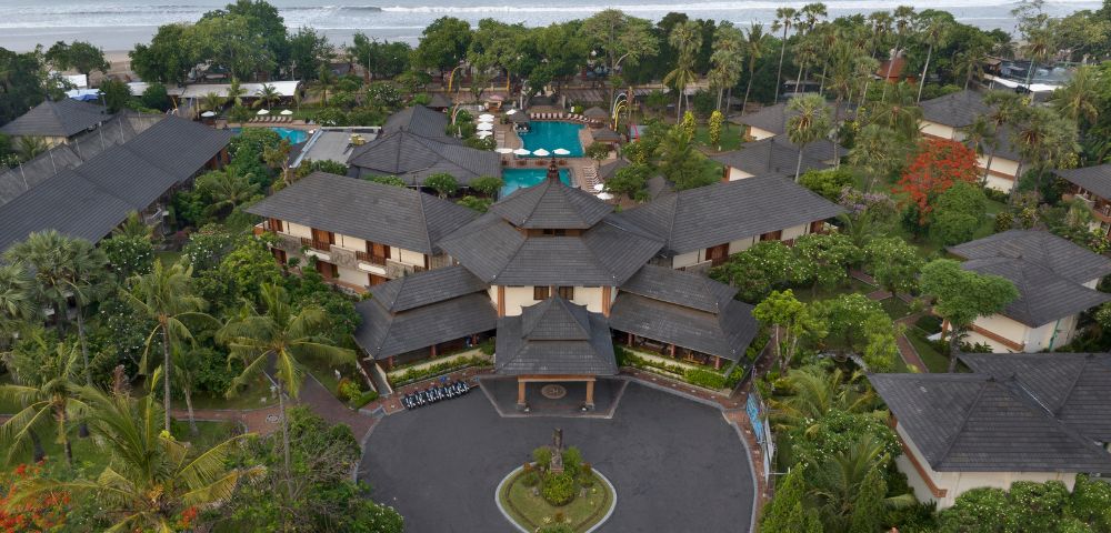 Aerial view of a tropical resort with lush gardens, a central circular driveway, multiple buildings, and a beach with waves in the background.