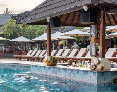 Poolside bar scene with a tiled counter, various drinks, and fresh coconuts. Behind are sun loungers and umbrellas, creating a relaxed, tropical vibe.