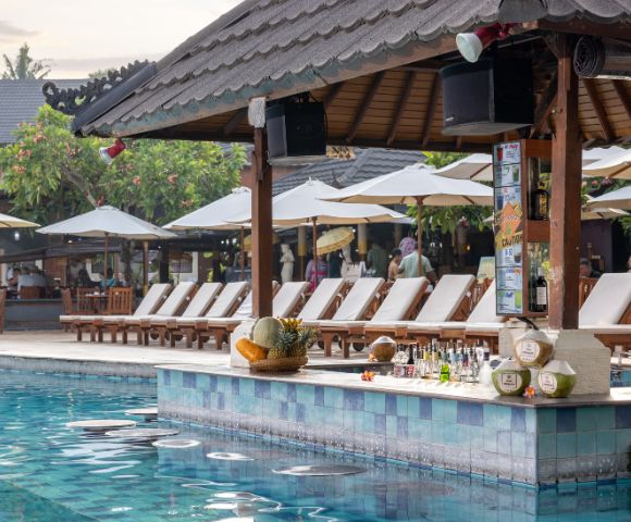 Poolside bar scene with a tiled counter, various drinks, and fresh coconuts. Behind are sun loungers and umbrellas, creating a relaxed, tropical vibe.