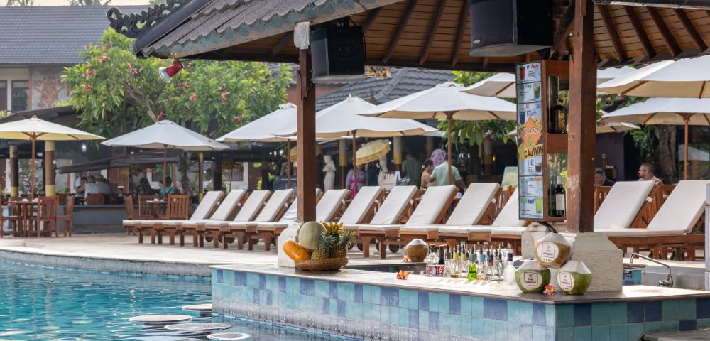 Poolside bar scene with a tiled counter, various drinks, and fresh coconuts. Behind are sun loungers and umbrellas, creating a relaxed, tropical vibe.