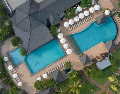 Aerial view of a resort with two pools surrounded by lounge chairs and white umbrellas, bordered by lush greenery and paved walkways. Calm and inviting atmosphere.