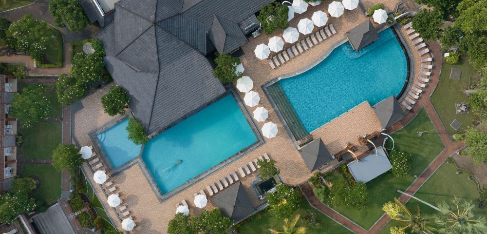 Aerial view of a resort with two pools surrounded by lounge chairs and white umbrellas, bordered by lush greenery and paved walkways. Calm and inviting atmosphere.