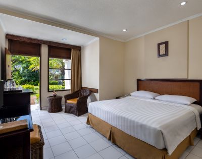 Bright hotel room with a neatly made double bed, wicker chair, and a view of lush greenery through large windows. Warm, inviting atmosphere.