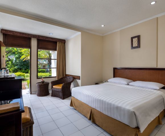 Bright hotel room with a neatly made double bed, wicker chair, and a view of lush greenery through large windows. Warm, inviting atmosphere.