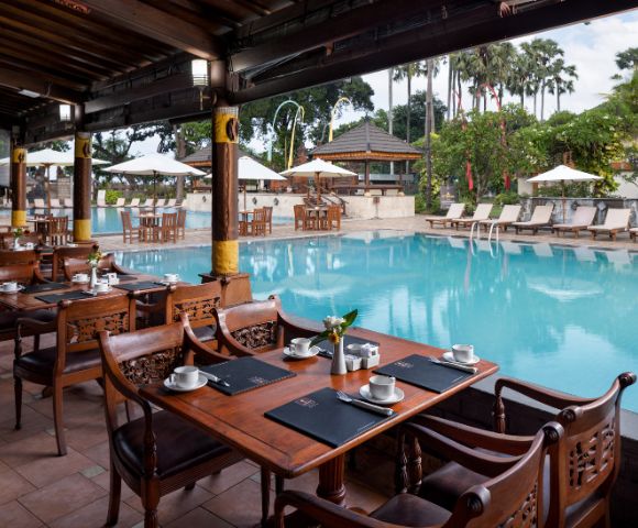 Outdoor restaurant beside a serene pool, featuring elegant wooden tables set with cups and placemats. Lush greenery and palm trees create a tropical ambiance.