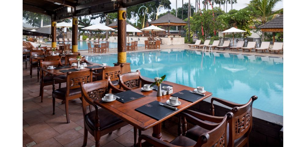 Outdoor restaurant beside a serene pool, featuring elegant wooden tables set with cups and placemats. Lush greenery and palm trees create a tropical ambiance.