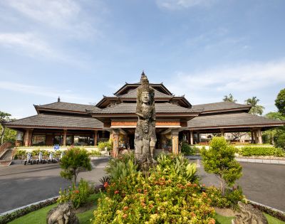 Traditional Balinese architecture with a tiered roof and wooden structure. A stone statue stands in front, surrounded by lush greenery under a clear sky.