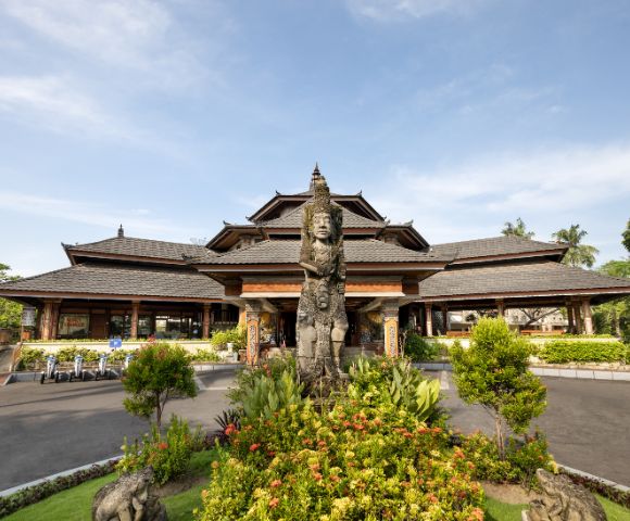 Traditional Balinese architecture with a tiered roof and wooden structure. A stone statue stands in front, surrounded by lush greenery under a clear sky.