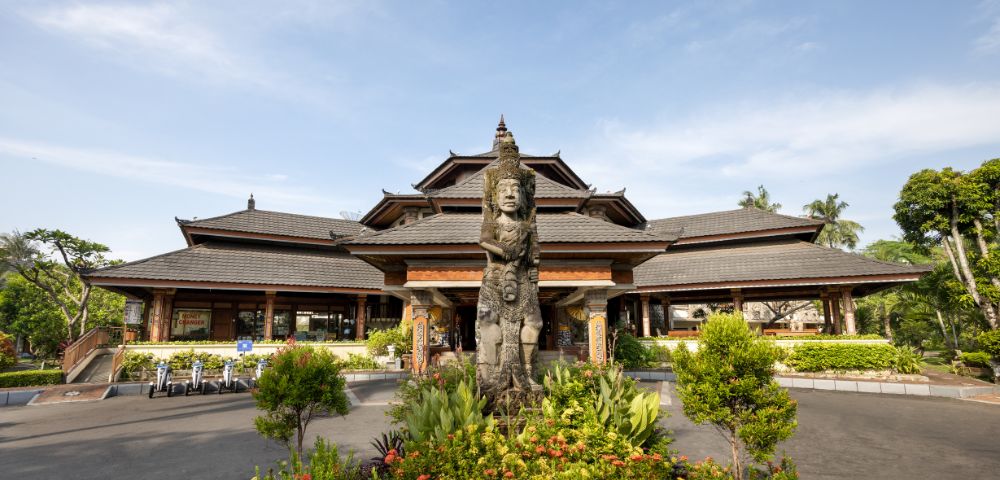 Traditional Balinese architecture with a tiered roof and wooden structure. A stone statue stands in front, surrounded by lush greenery under a clear sky.