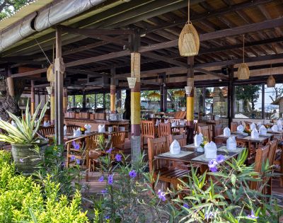 Open-air wooden restaurant with neatly set tables, white napkins, and hanging lanterns. Surrounded by lush greenery and vibrant purple flowers, exuding a serene, tropical vibe.