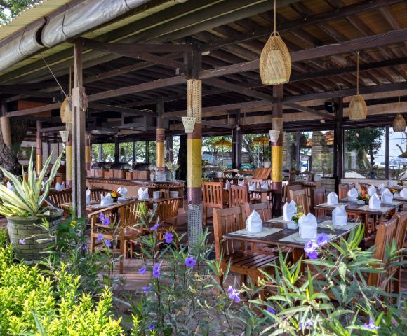 Open-air wooden restaurant with neatly set tables, white napkins, and hanging lanterns. Surrounded by lush greenery and vibrant purple flowers, exuding a serene, tropical vibe.