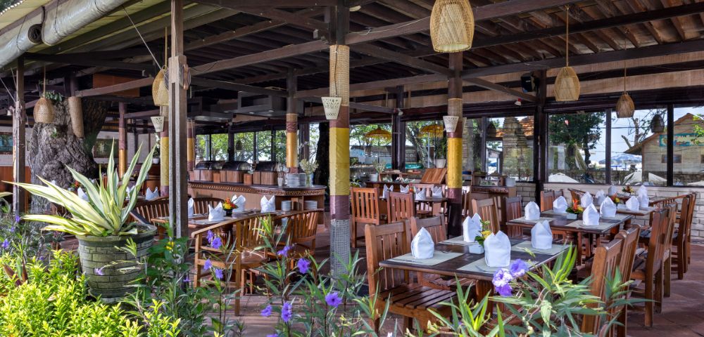 Open-air wooden restaurant with neatly set tables, white napkins, and hanging lanterns. Surrounded by lush greenery and vibrant purple flowers, exuding a serene, tropical vibe.