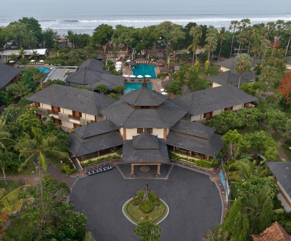 Aerial view of a tropical resort with lush gardens, a central circular driveway, multiple buildings, and a beach with waves in the background.