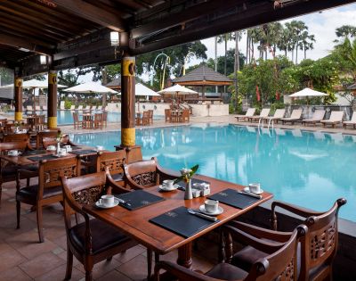 Outdoor restaurant beside a serene pool, featuring elegant wooden tables set with cups and placemats. Lush greenery and palm trees create a tropical ambiance.