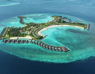 Aerial view of a tropical island resort surrounded by turquoise waters. Overwater bungalows extend into the sea, and lush greenery covers the island.