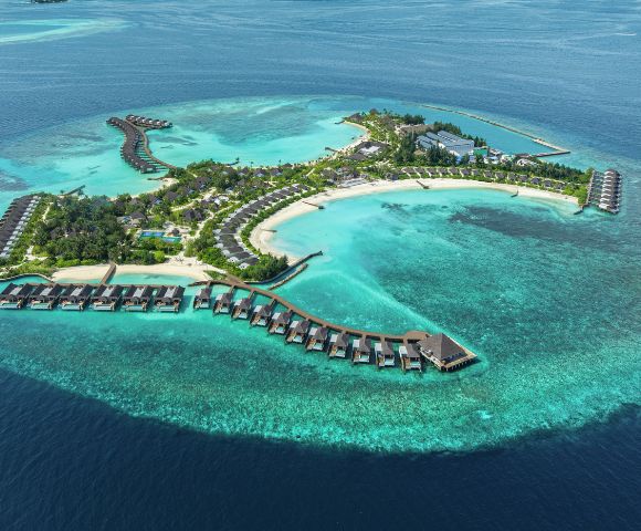 Aerial view of a tropical island resort surrounded by turquoise waters. Overwater bungalows extend into the sea, and lush greenery covers the island.