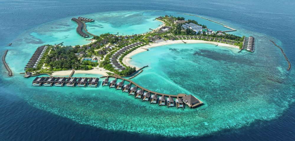 Aerial view of a tropical island resort surrounded by turquoise waters. Overwater bungalows extend into the sea, and lush greenery covers the island.