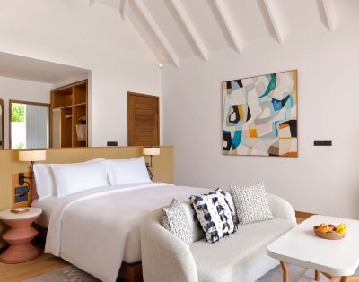 Modern hotel room with a plush white bed, abstract painting, and cozy sofa. Warm wood accents, geometric lamps, and a bowl of fruit create a serene ambiance.