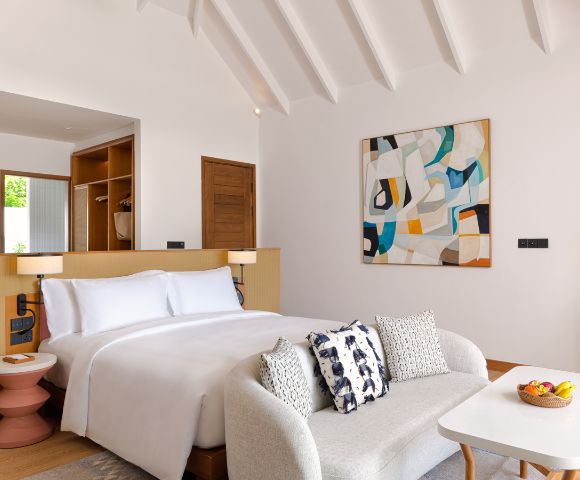 Modern hotel room with a plush white bed, abstract painting, and cozy sofa. Warm wood accents, geometric lamps, and a bowl of fruit create a serene ambiance.