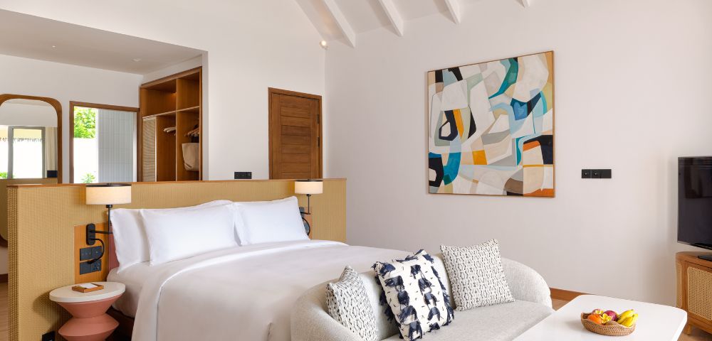 Modern hotel room with a plush white bed, abstract painting, and cozy sofa. Warm wood accents, geometric lamps, and a bowl of fruit create a serene ambiance.