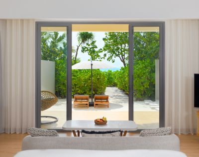 Modern hotel room with a view through glass doors of a lush green garden, sun loungers, and a white parasol. Cozy indoor seating creates a peaceful, inviting atmosphere.