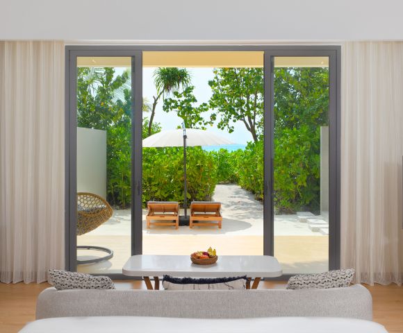 Modern hotel room with a view through glass doors of a lush green garden, sun loungers, and a white parasol. Cozy indoor seating creates a peaceful, inviting atmosphere.