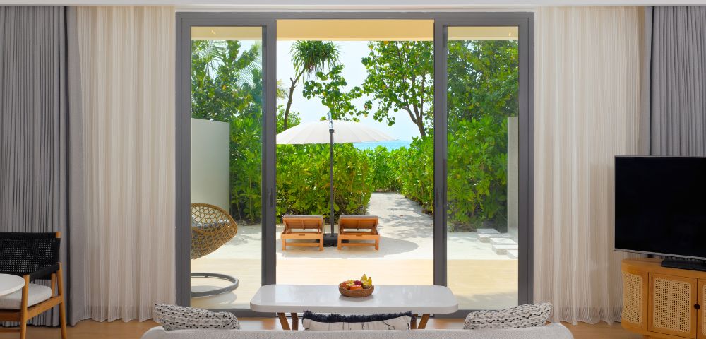 Modern hotel room with a view through glass doors of a lush green garden, sun loungers, and a white parasol. Cozy indoor seating creates a peaceful, inviting atmosphere.