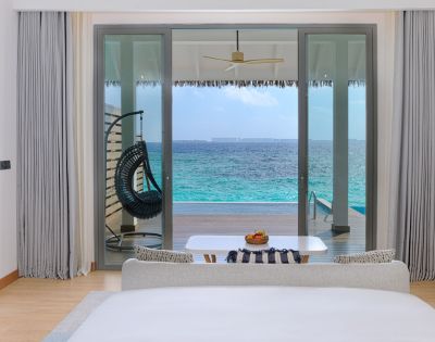 Elegant beachfront room with glass doors opening to a turquoise ocean. A hanging chair, table with fruit, and serene decor create a tranquil ambiance.