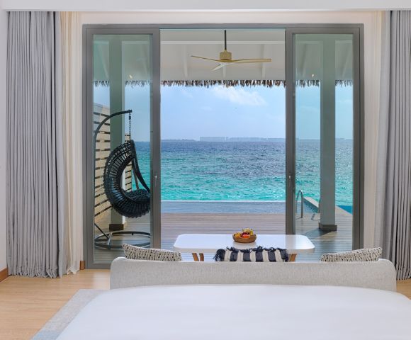 Elegant beachfront room with glass doors opening to a turquoise ocean. A hanging chair, table with fruit, and serene decor create a tranquil ambiance.