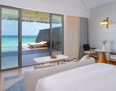 Luxurious beachside room with modern decor, white linens, and glass doors opening to a turquoise ocean view. Relaxing, serene ambiance.