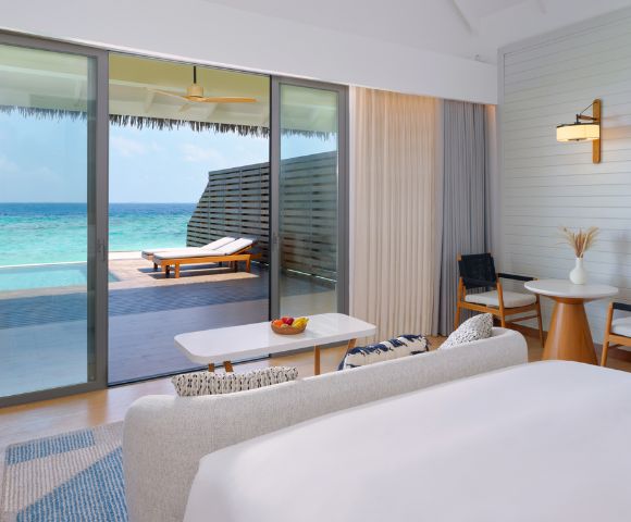 Luxurious beachside room with modern decor, white linens, and glass doors opening to a turquoise ocean view. Relaxing, serene ambiance.