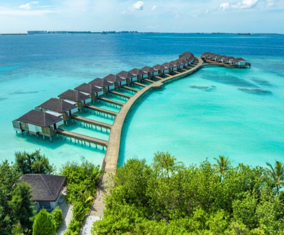 A series of overwater bungalows on a curved wooden walkway stretch into crystal-clear turquoise ocean, surrounded by lush green foliage under a bright sky.