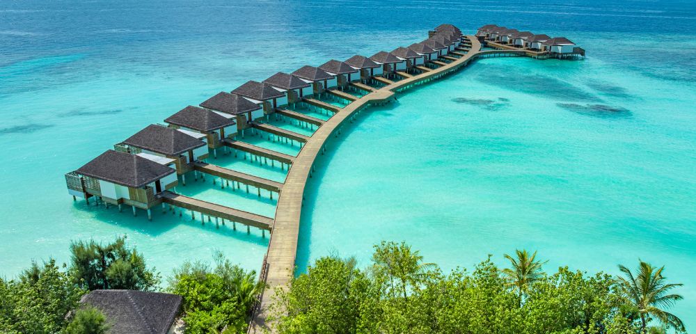 A series of overwater bungalows on a curved wooden walkway stretch into crystal-clear turquoise ocean, surrounded by lush green foliage under a bright sky.
