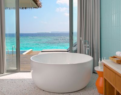 Luxurious bathroom with a round white tub facing ocean view. Light gray curtains frame a glass door leading to a wooden deck. Calm, serene ambiance.