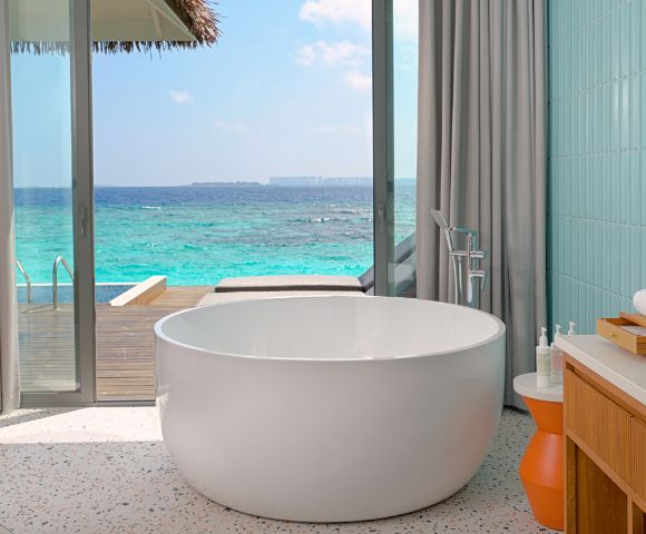Luxurious bathroom with a round white tub facing ocean view. Light gray curtains frame a glass door leading to a wooden deck. Calm, serene ambiance.