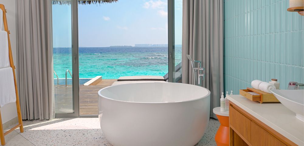 Luxurious bathroom with a round white tub facing ocean view. Light gray curtains frame a glass door leading to a wooden deck. Calm, serene ambiance.